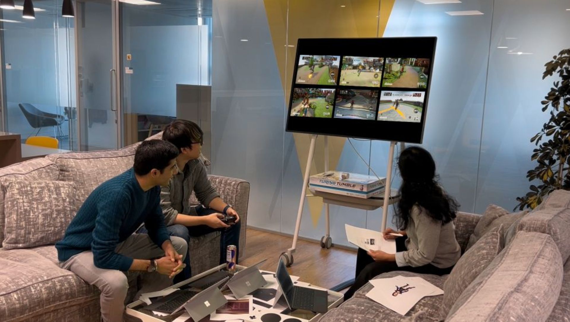Three Microsoft researchers in a lounge — one holding a game controller — watching a large display showing a grid of six Bleeding Edge gameplay clips, with laptops and prototype hardware spread across the table in front of them.