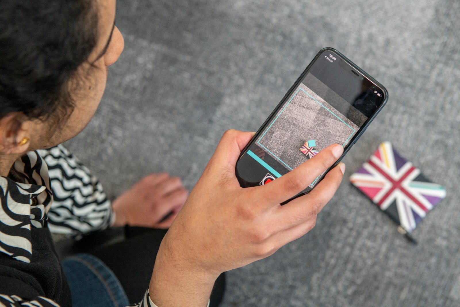 Over-the-shoulder view of a woman holding a phone running Find My Things. The screen shows the live camera view of the carpet with a detection box drawn around a small Union Jack coaster, which is lying on the floor a short distance in front of her.